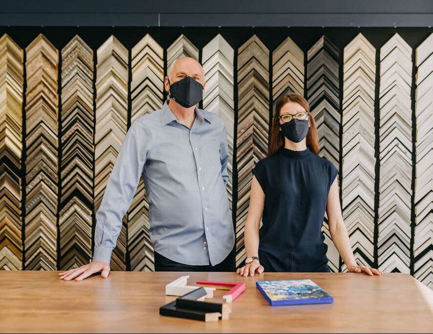 Pete and Laura, owners of Bergeron Art + Frames posing for a picture in front of their work desk